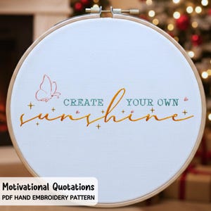 以下が含まれることがあります： 白い布の刺繍枠に、「CREATE YOUR OWN sunshine」という言葉が金と緑の糸で刺繍されています。ピンクの蝶と小さな十字形のアクセントも刺繍されています。木製の枠は金属製の留め金で固定されています。