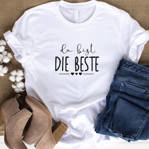 K&ouml;nnte beinhalten: Wei&szlig;es T-Shirt mit schwarzem Text, der "du bist die beste" mit drei kleinen Herzen darunter lautet. Das Shirt liegt flach auf einer braunen Oberfl&auml;che mit einem Paar braunen Stiefeln und einer blauen Jeans.