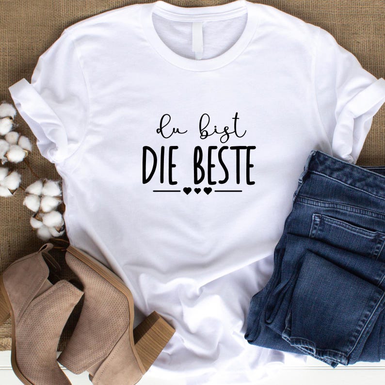 K&ouml;nnte beinhalten: Wei&szlig;es T-Shirt mit schwarzem Text, der "du bist die beste" mit drei kleinen Herzen darunter lautet. Das Shirt liegt flach auf einer braunen Oberfl&auml;che mit einem Paar braunen Stiefeln und einer blauen Jeans.