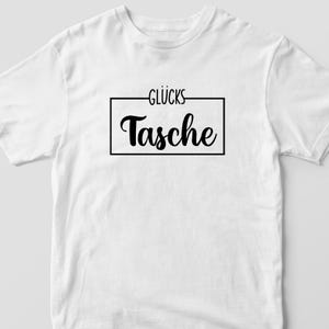 Op de afbeelding: Wit T-shirt met zwarte tekst die "Gl&uuml;cks Tasche" binnen een zwart rechthoek leest.
