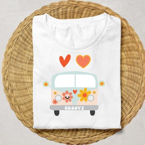 Puede incluir: Camiseta blanca con una ilustraci&oacute;n de dibujos animados de una furgoneta retro, flores y la palabra "GROOVY". Dos corazones est&aacute;n encima de la furgoneta. La camiseta est&aacute; doblada sobre una superficie de mimbre tejido.