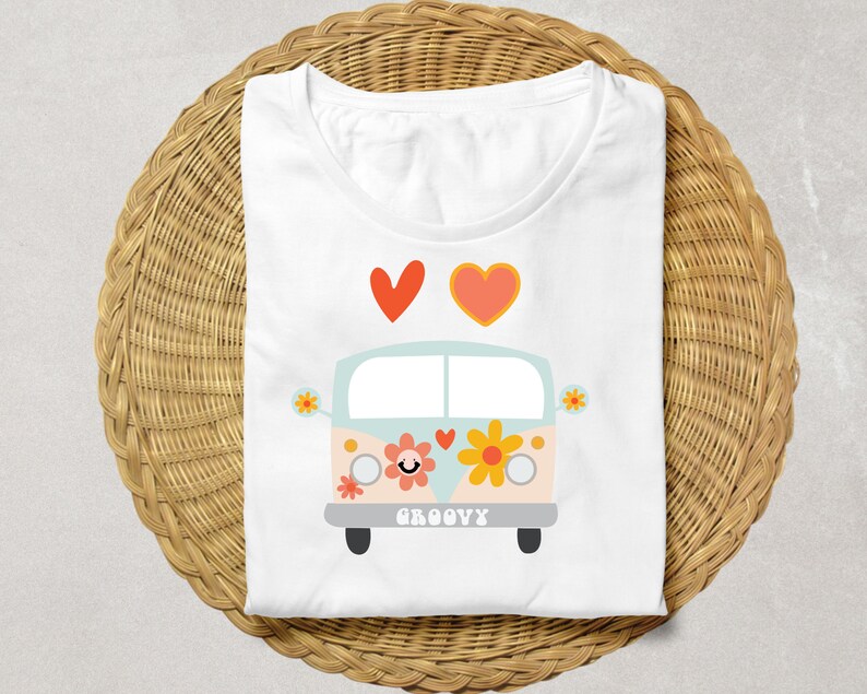 Puede incluir: Camiseta blanca con una ilustraci&oacute;n de dibujos animados de una furgoneta retro, flores y la palabra "GROOVY". Dos corazones est&aacute;n encima de la furgoneta. La camiseta est&aacute; doblada sobre una superficie de mimbre tejido.