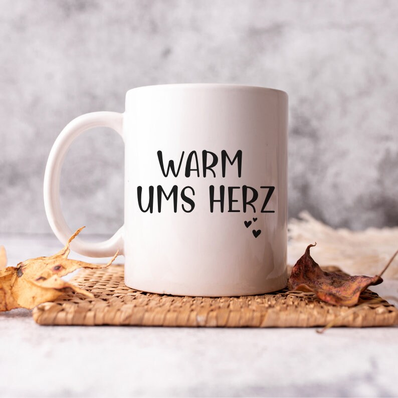 May include: White ceramic mug with black text that reads "Warm ums Herz" and three small black hearts. The mug is sitting on a woven wicker coaster.
