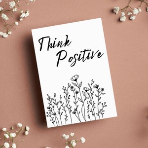 Op de afbeelding: Een witte kaart met zwarte tekst die "Think Positive" zegt en een zwarte lijntekening van een veld met wilde bloemen.