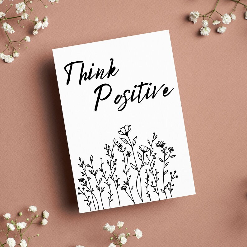 Op de afbeelding: Een witte kaart met zwarte tekst die "Think Positive" zegt en een zwarte lijntekening van een veld met wilde bloemen.