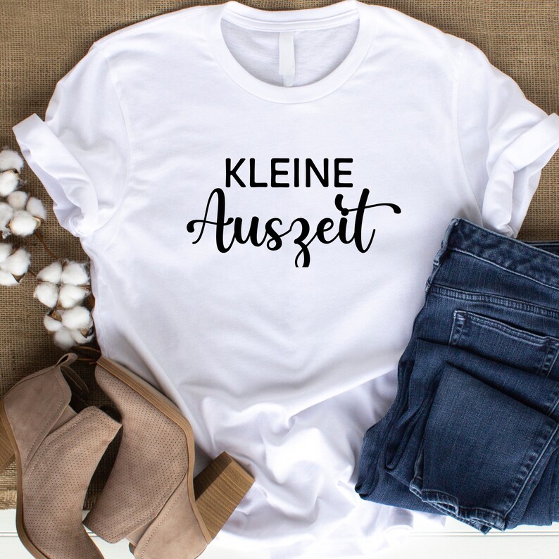 May include: White t-shirt with black text that reads "KLEINE Auszeit". The shirt is laid flat on a brown surface with a pair of blue jeans and brown ankle boots.