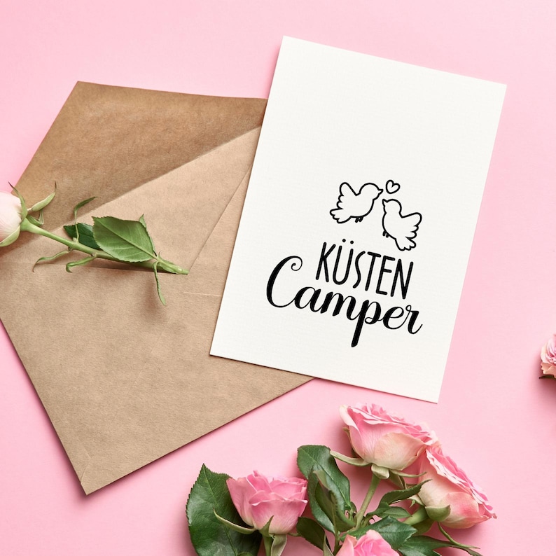 May include: A white card with black text and a simple illustration of two birds facing each other. The text reads "K&uuml;sten Camper". The card is on a pink background with a brown envelope and pink roses.
