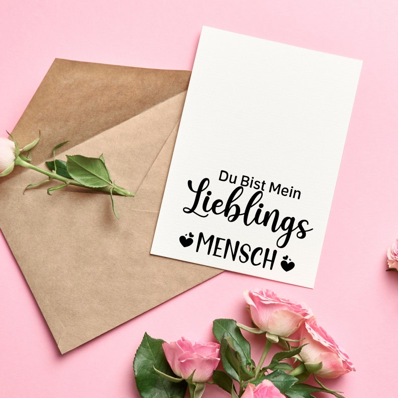 May include: A white card with the text "Du Bist Mein Lieblings Mensch" in black lettering with two small hearts on either side of the word "Mensch". The card is on a pink background with a brown envelope and pink roses.