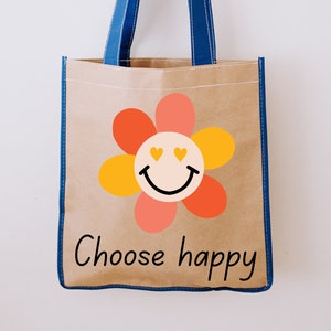 Puede incluir: Un bolso tote marr&oacute;n con asas y ribetes azules. El bolso presenta un gr&aacute;fico de flores de colores con una carita sonriente y las palabras "Choose happy" en letras negras. El bolso est&aacute; hecho de un material resistente.