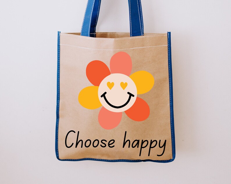 Puede incluir: Un bolso tote marr&oacute;n con asas y ribetes azules. El bolso presenta un gr&aacute;fico de flores de colores con una carita sonriente y las palabras "Choose happy" en letras negras. El bolso est&aacute; hecho de un material resistente.