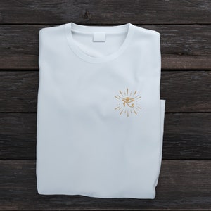 May include: White t-shirt with a gold eye of Horus graphic design on the front.