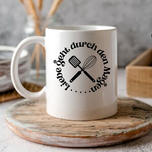 May include: White ceramic coffee mug with a black graphic of crossed kitchen utensils and the text "Liebe geht durch den Magen".