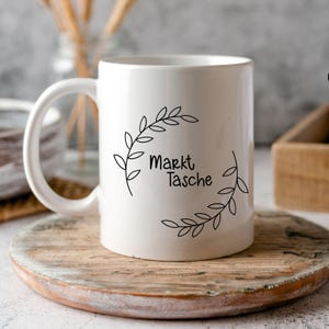 Op de afbeelding: Witte keramische mok met een zwart ontwerp van een krans van bladeren en de tekst "Markt Tasche".