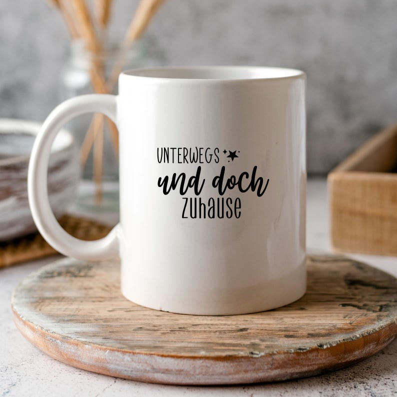 May include: White ceramic mug with black text that reads "Unterwegs und doch zuhause" which translates to "On the way and yet at home".