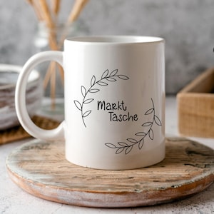 K&ouml;nnte beinhalten: Wei&szlig;e Keramiktasse mit einem schwarzen Design aus einem Kranz aus Bl&auml;ttern und dem Text "Markt Tasche".