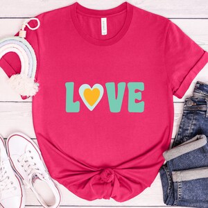 Puede incluir: Una camiseta magenta con la palabra "LOVE" en letras turquesas y un coraz&oacute;n amarillo. La camiseta se combina con vaqueros azules y zapatillas blancas. Un arco&iacute;ris decorativo tambi&eacute;n est&aacute; en la imagen.