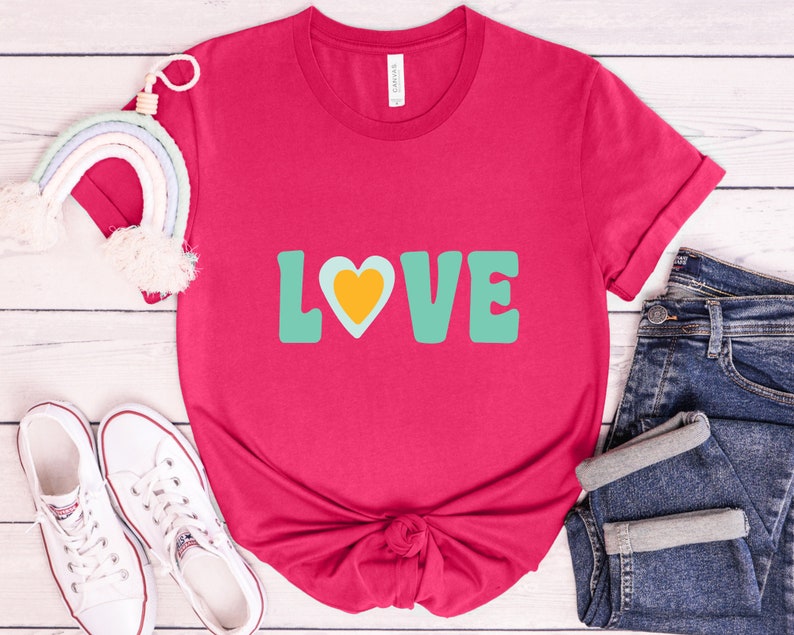 Puede incluir: Una camiseta magenta con la palabra "LOVE" en letras turquesas y un coraz&oacute;n amarillo. La camiseta se combina con vaqueros azules y zapatillas blancas. Un arco&iacute;ris decorativo tambi&eacute;n est&aacute; en la imagen.
