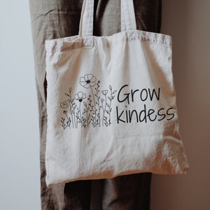 Op de afbeelding: Een witte canvas tote bag met zwarte bloemenillustraties en de tekst "Grow kindness".