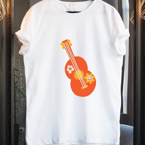 Puede incluir: Camiseta blanca con un dise&ntilde;o de guitarra de dibujos animados en naranja, amarillo y blanco. La guitarra tiene una flor y una cara de flor sonriente en el cuerpo. La camiseta est&aacute; colgada de una percha negra.