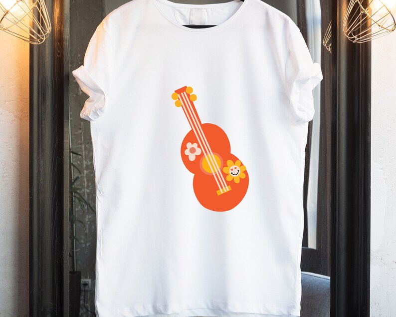 Puede incluir: Camiseta blanca con un dise&ntilde;o de guitarra de dibujos animados en naranja, amarillo y blanco. La guitarra tiene una flor y una cara de flor sonriente en el cuerpo. La camiseta est&aacute; colgada de una percha negra.