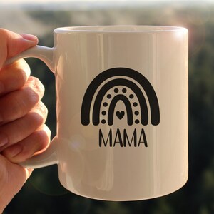 Pu&ograve; includere: Una tazza in ceramica bianca con un design arcobaleno bianco e nero e la parola "MAMA" stampata sotto.