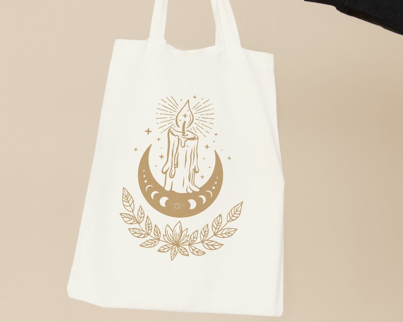 May include: A white tote bag with a gold design of a candle and a crescent moon with stars. The design includes a stylized candle with a flame and a crescent moon with phases.