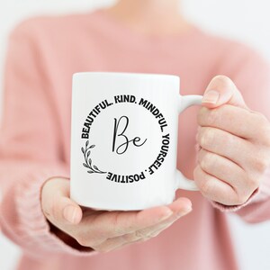 May include: White ceramic mug with a black circle design that says "Be Beautiful. Kind. Mindful. Positive. Yourself."  The mug is being held by a person's hands.