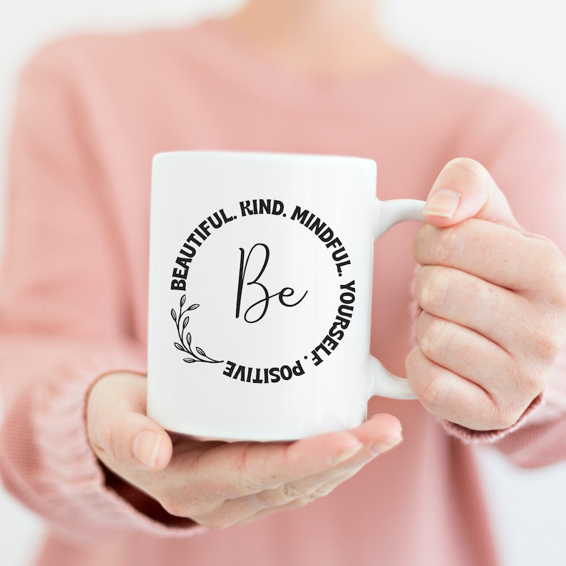 May include: White ceramic mug with a black circle design that says "Be Beautiful. Kind. Mindful. Positive. Yourself."  The mug is being held by a person's hands.