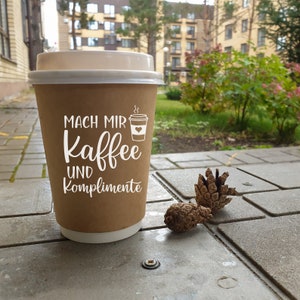 May include: A brown paper coffee cup with a white lid. The cup has the text "Mach mir Kaffee und Komplimente" printed on it. The cup is sitting on a gray sidewalk.
