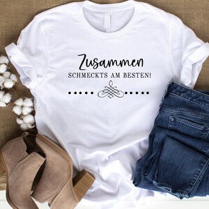 May include: White t-shirt with the text "Zusammen schmeckt's am besten!" in black lettering. The shirt is folded and displayed on a brown burlap background with a pair of brown ankle boots and blue jeans.