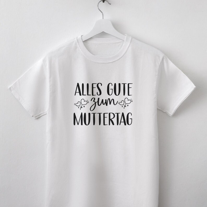 Pu&ograve; includere: Maglietta bianca con testo nero che dice "Alles Gute zum Muttertag" con piccoli cuori intorno alla parola "zum".