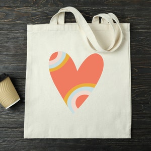 Puede incluir: Un bolso tote de color crema con un gran dise&ntilde;o de coraz&oacute;n coral. El coraz&oacute;n presenta detalles de arco&iacute;ris en amarillo, azul y rosa. El bolso tiene dos asas de color crema y est&aacute; hecho de lona resistente. A la izquierda del bolso hay una taza de caf&eacute; desechable.