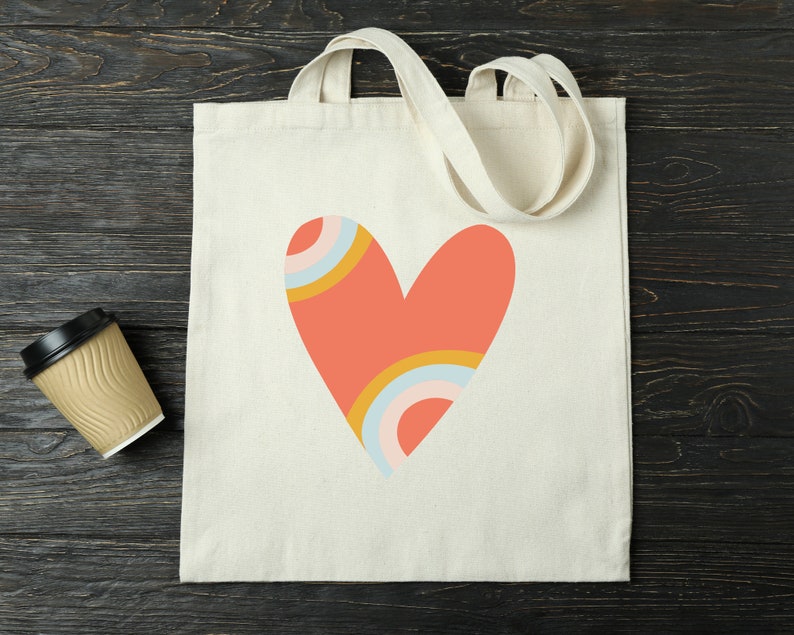 Puede incluir: Un bolso tote de color crema con un gran dise&ntilde;o de coraz&oacute;n coral. El coraz&oacute;n presenta detalles de arco&iacute;ris en amarillo, azul y rosa. El bolso tiene dos asas de color crema y est&aacute; hecho de lona resistente. A la izquierda del bolso hay una taza de caf&eacute; desechable.