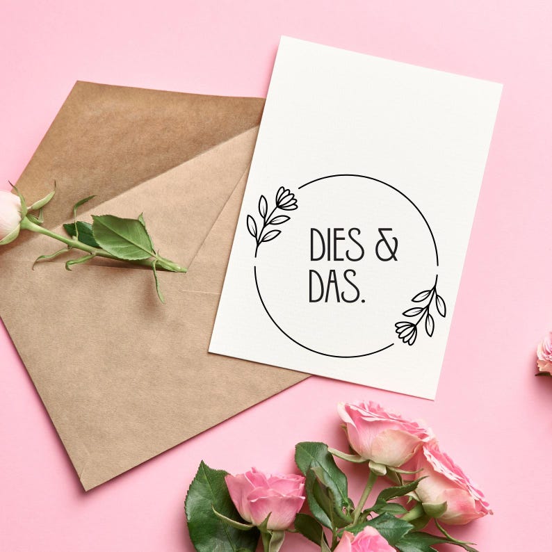 Op de afbeelding: Een witte kaart met een zwarte cirkel en de tekst "Dies & Das." De cirkel is versierd met twee kleine bloemmotieven. De kaart ligt op een roze achtergrond met een bruine envelop en roze rozen.