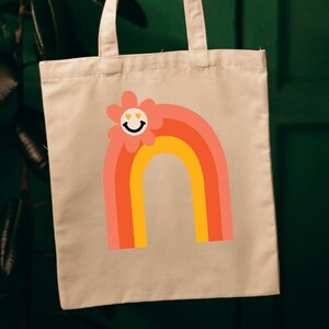 Puede incluir: Bolso tote beige con un dise&ntilde;o de arco&iacute;ris colorido y una flor sonriente. El arco&iacute;ris tiene capas de naranja, coral y amarillo. La flor tiene una cara sonriente con ojos en forma de coraz&oacute;n. La bolsa cuelga sobre un fondo verde oscuro.