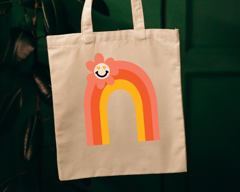 Puede incluir: Bolso tote beige con un dise&ntilde;o de arco&iacute;ris colorido y una flor sonriente. El arco&iacute;ris tiene capas de naranja, coral y amarillo. La flor tiene una cara sonriente con ojos en forma de coraz&oacute;n. La bolsa cuelga sobre un fondo verde oscuro.
