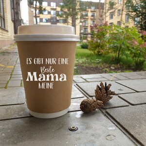 Pu&ograve; includere: Un bicchiere di carta marrone con un coperchio bianco. Il bicchiere ha la scritta "Es gibt nur eine Beste Mama Meine" stampata su di esso. Il bicchiere &egrave; appoggiato su una superficie di cemento grigia con due piccole pigne accanto.