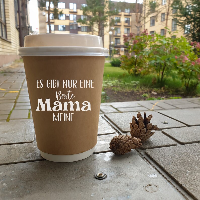 Pu&ograve; includere: Un bicchiere di carta marrone con un coperchio bianco. Il bicchiere ha la scritta "Es gibt nur eine Beste Mama Meine" stampata su di esso. Il bicchiere &egrave; appoggiato su una superficie di cemento grigia con due piccole pigne accanto.