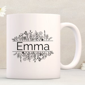 Op de afbeelding: Witte keramische mok met een zwarte omtrek van bloemen en de naam "Emma" gedrukt op de voorkant.