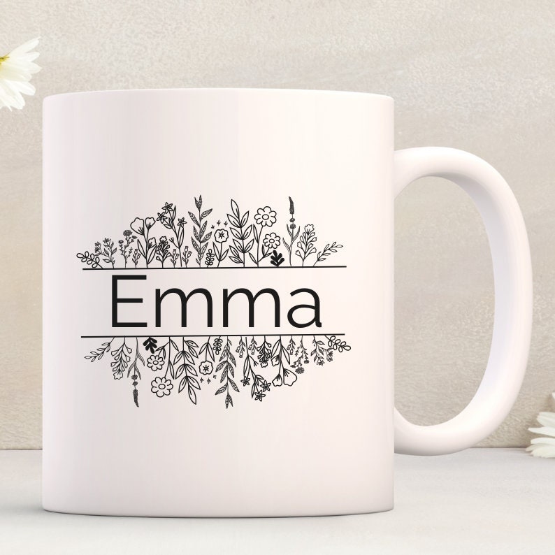 Op de afbeelding: Witte keramische mok met een zwarte omtrek van bloemen en de naam "Emma" gedrukt op de voorkant.