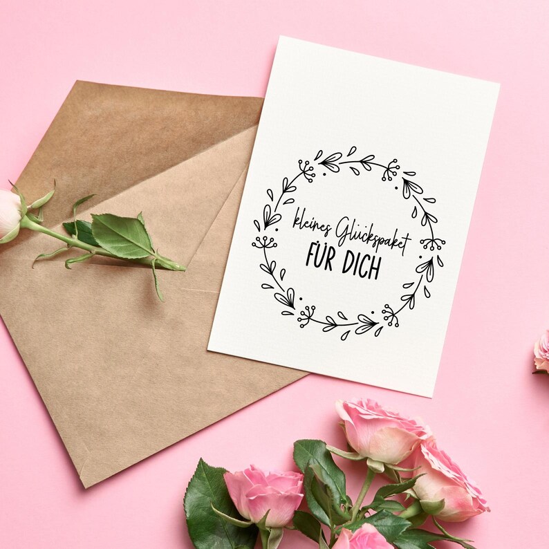 K&ouml;nnte beinhalten: Eine wei&szlig;e Karte mit einem schwarzen Blumenkranz und dem Text "Kleines Gl&uuml;ckspacket f&uuml;r Dich" auf rosa Hintergrund mit rosa Rosen.