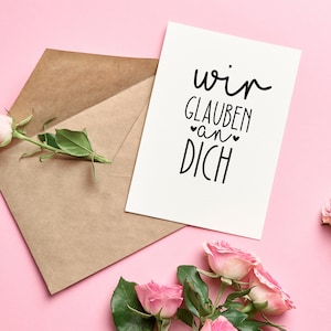 Peut inclure: Une carte blanche avec le texte "wir glauben an dich" en lettres noires, avec deux petits c&oelig;urs autour du mot "an". La carte est sur un fond rose avec une enveloppe brune et des roses roses.