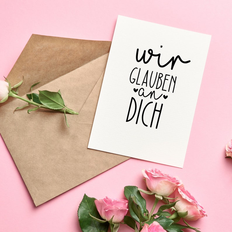 Peut inclure: Une carte blanche avec le texte "wir glauben an dich" en lettres noires, avec deux petits c&oelig;urs autour du mot "an". La carte est sur un fond rose avec une enveloppe brune et des roses roses.