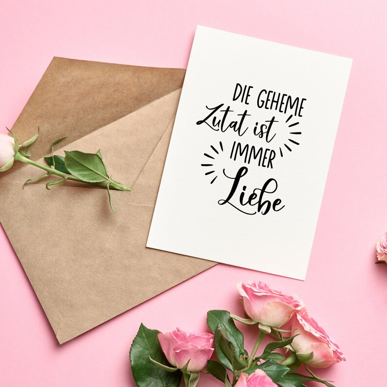 May include: A white card with the text "DIE GEHEIME Zutat ist IMMER Liebe" in black cursive lettering. The card is on a pink background with a brown envelope and pink roses.