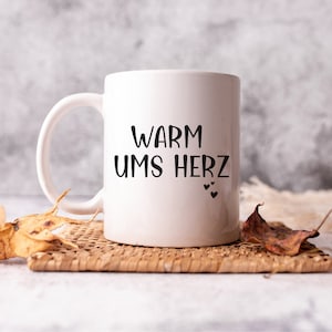 May include: White ceramic mug with black text that reads "Warm ums Herz" and three small black hearts. The mug is sitting on a woven wicker coaster.