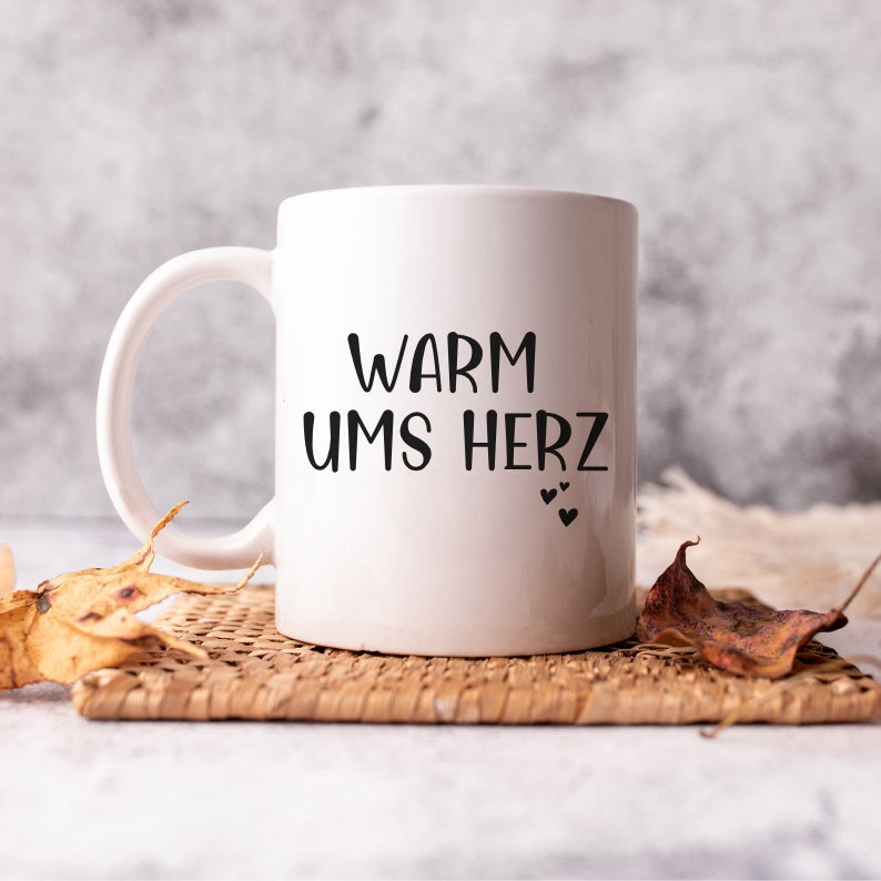 May include: White ceramic mug with black text that reads "Warm ums Herz" and three small black hearts. The mug is sitting on a woven wicker coaster.
