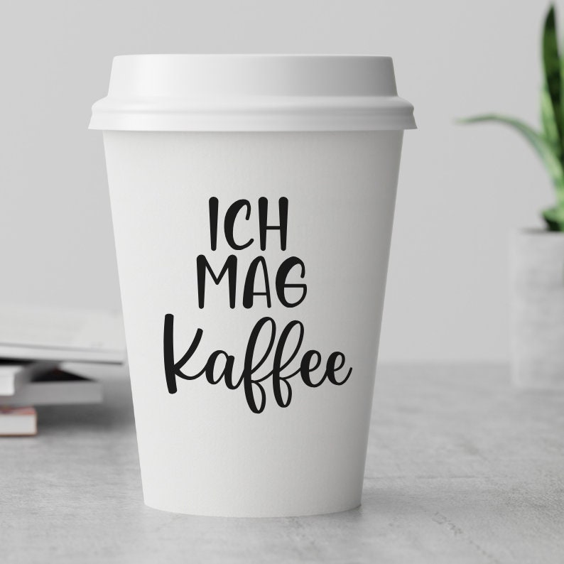 May include: White to-go coffee cup with black text that reads "Ich mag Kaffee" which translates to "I like coffee" in English.