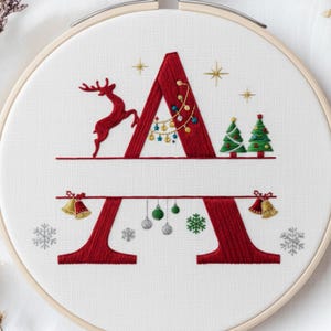 May include: Embroidered Christmas-themed hoop art featuring a red letter "A" with festive details. A reindeer, Christmas trees, bells, and ornaments adorn the design. The embroidery is set against a white fabric background, framed by a wooden hoop.