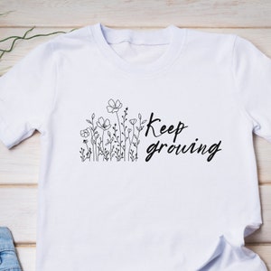 Op de afbeelding: Wit T-shirt met een zwarte lijntekening van bloemen en de tekst "Keep growing" erop.