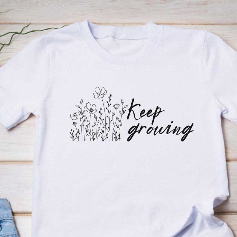 Op de afbeelding: Wit T-shirt met een zwarte lijntekening van bloemen en de tekst "Keep growing" erop.